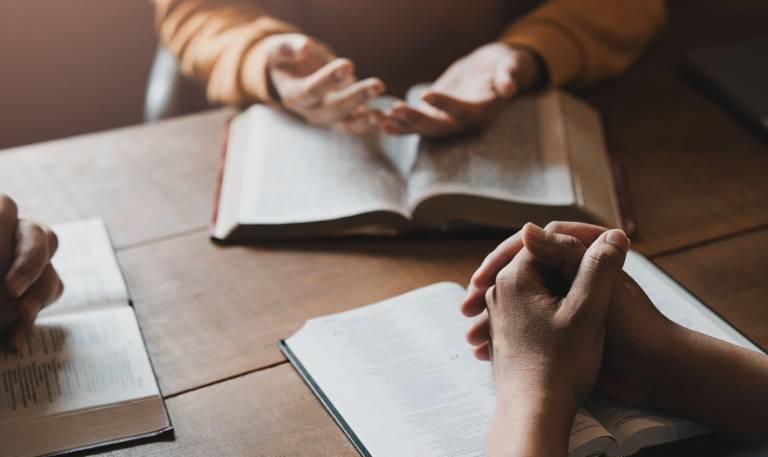 A group of Christians sit together and pray around a wooden table with blurred open Bible pages